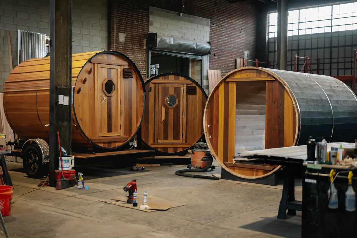 Three barrel saunas in the Nomad workshop showing natural cedar, mixed wood tones, and Yakisugi charred exterior finishes